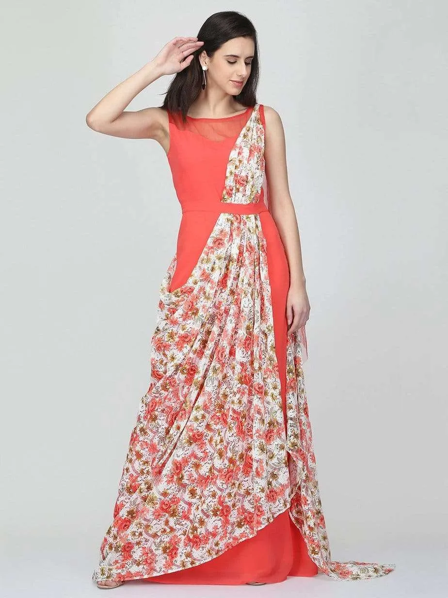 A woman in the Eavan Coral Drape Gown with a floral printed overlay poses against a plain light background, gently touching her hair and looking down with a subtle smile.
